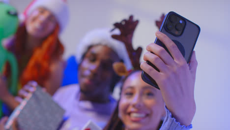 Studioaufnahme-Von-Freunden-Der-Generation-Z-Zu-Weihnachten,-Die-Mit-Weihnachtsmütze-Und-Rentiergeweih-Auf-Dem-Sofa-Sitzen-Und-Ein-Selfie-Mit-Dem-Mobiltelefon-Machen-2